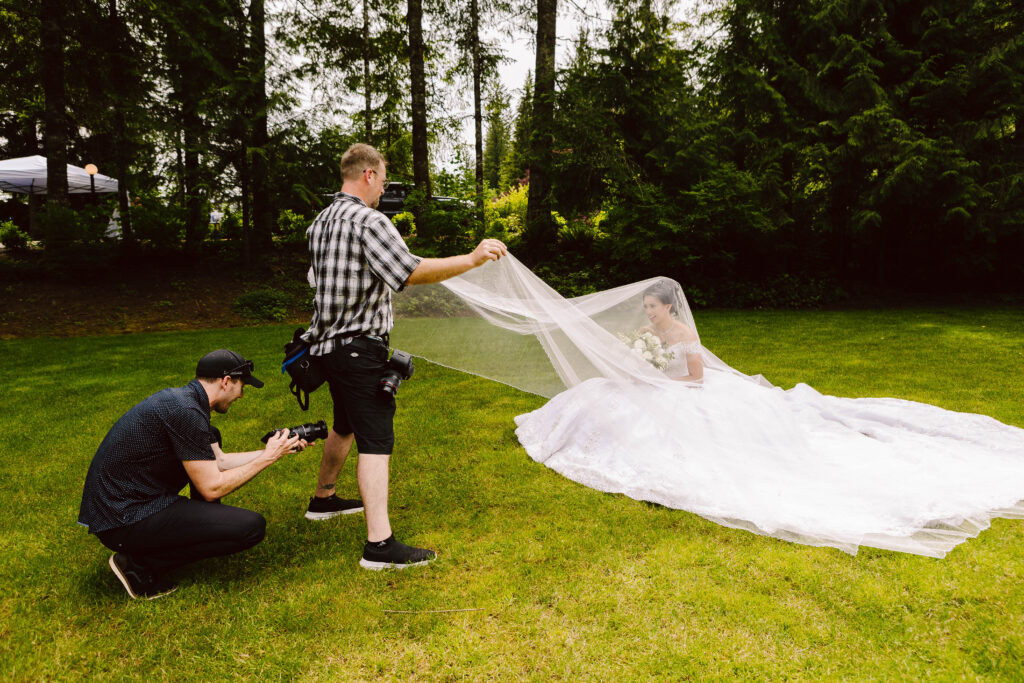 Snohomish Wedding Photographer GSquared Weddings A talented photographer kneels to capture a stunning image of a bride in her white dress sitting gracefully on the grass. Meanwhile, an assistant meticulously adjusts her veil. They find themselves enveloped by natures beauty, with tall trees and lush greenery all around. Serving Seattle, Snohomish and Washington State