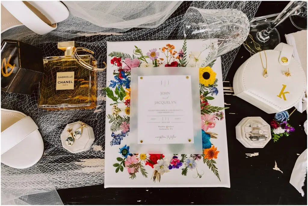 Snohomish Wedding Photographer GSquared Weddings A flat lay image showcases wedding invitations amidst a vibrant floral design, alongside white shoes, Chanel perfume, jewelry, and rings in a box on dark machias. Nearby rests a heart-shaped case with a K, all beautifully arranged on tulle. Serving Seattle, Snohomish and Washington State