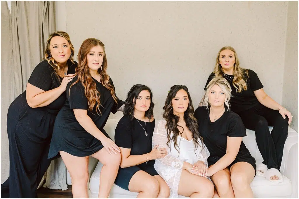 Snohomish Wedding Photographer GSquared Weddings A group of six women poses indoors in Machias. The woman in the center dons a sheer white robe, while the others wear black outfits. Arranged around a white sofa, they look at the camera with serious expressions, capturing a striking contrast and an aura of elegance. Serving Seattle, Snohomish and Washington State