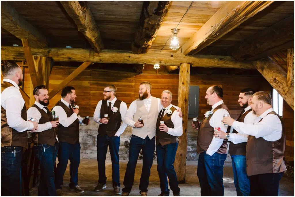 Snohomish Wedding Photographer GSquared Weddings A group of eight men in vests and jeans are standing in a rustic wooden room in Machias, smiling and holding drink bottles. They appear to be at a casual gathering or celebration, possibly a wedding. The mood is relaxed and joyful. Serving Seattle, Snohomish and Washington State