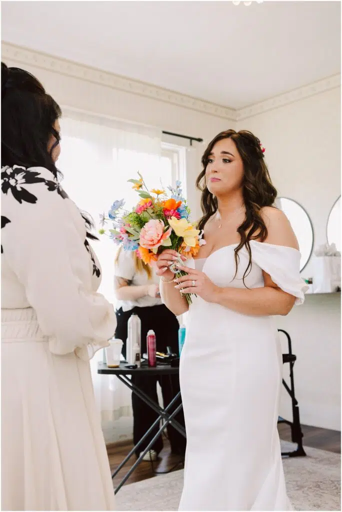 Snohomish Wedding Photographer GSquared Weddings In a charming Machias setting, a bride in an off-shoulder white dress holds a colorful bouquet, facing someone in a cream outfit. Indoors, with salon tools visible behind them, natural light streams through sheer curtains for an ethereal touch. Serving Seattle, Snohomish and Washington State
