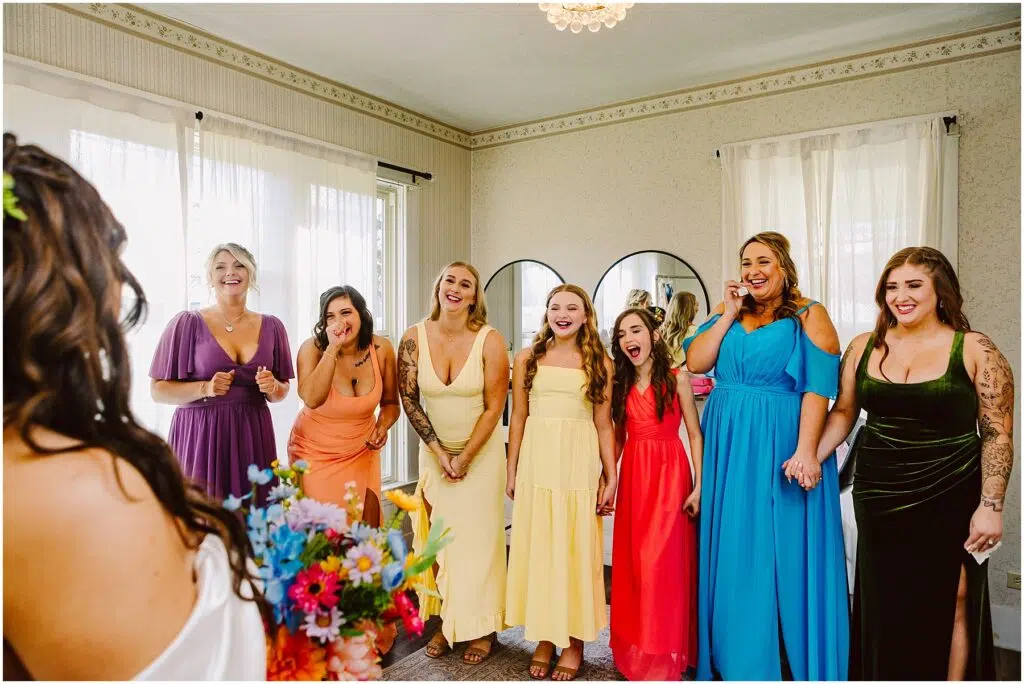 Snohomish Wedding Photographer GSquared Weddings A bride stands with her back to the camera holding a colorful bouquet. In front of her, a group of six bridesmaids and a flower girl from Machias react with joy and surprise. They are wearing dresses in various colors, and the room is decorated with mirrors and curtains. Serving Seattle, Snohomish and Washington State