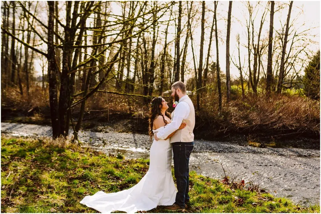 Snohomish Wedding Photographer GSquared Weddings A couple in wedding attire embraces by the scenic Machias River, surrounded by bare trees and greenery. The sun filters through the branches, creating a peaceful and romantic atmosphere. Serving Seattle, Snohomish and Washington State
