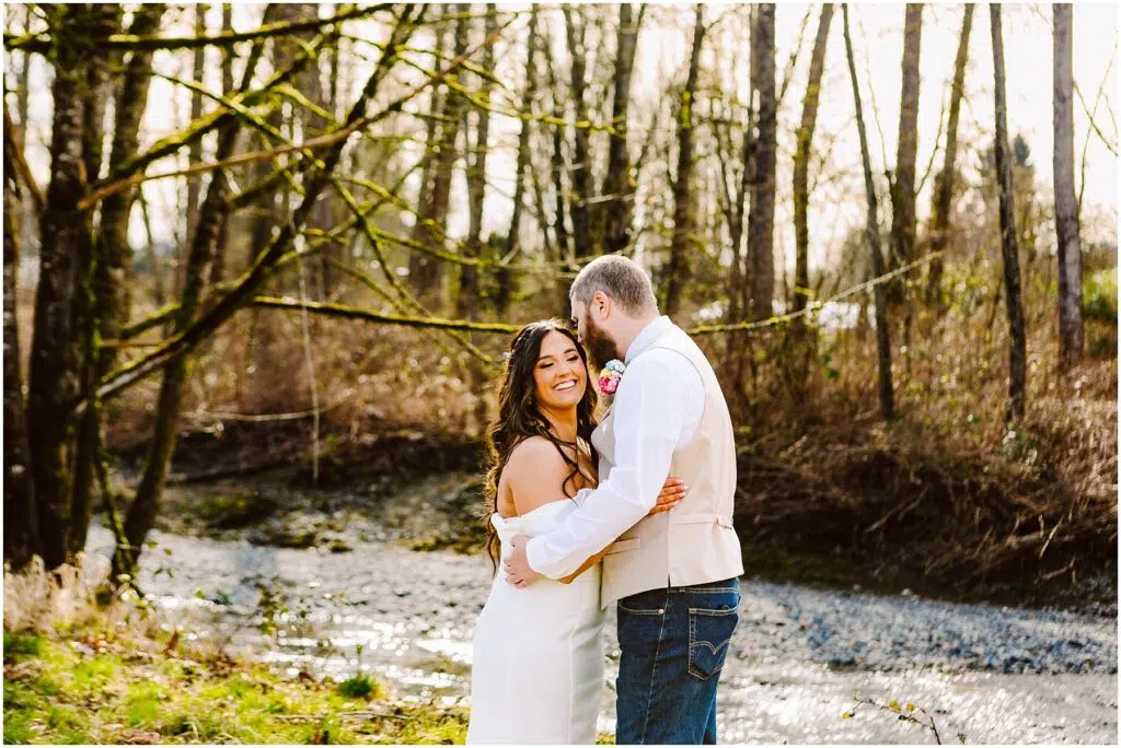 Snohomish Wedding Photographer GSquared Weddings A couple embraces near a small stream in a wooded area reminiscent of Machias. The woman, smiling in her white dress, is held gently by the man in a light shirt and jeans. Leafless trees frame their moment on this sunny day. Serving Seattle, Snohomish and Washington State