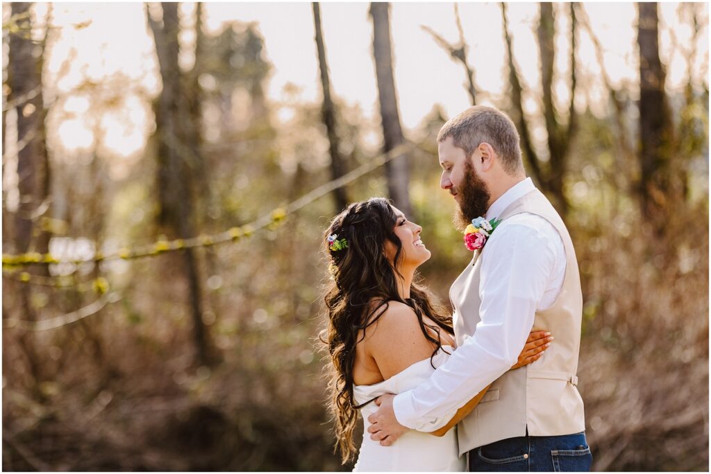 Snohomish Wedding Photographer GSquared Weddings In a serene Machias forest clearing, a couple embraces. The woman, adorned with long curly hair and a floral headpiece, wears a white dress. Beside her stands the man, bearded and donning a white shirt and vest with a colorful boutonniere. Sunlight filters through the trees behind them. Serving Seattle, Snohomish and Washington State