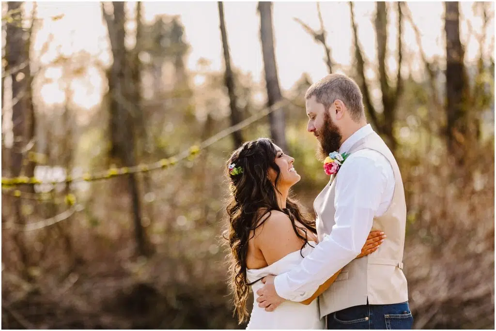 Snohomish Wedding Photographer GSquared Weddings In a serene Machias forest clearing, a couple embraces. The woman, adorned with long curly hair and a floral headpiece, wears a white dress. Beside her stands the man, bearded and donning a white shirt and vest with a colorful boutonniere. Sunlight filters through the trees behind them. Serving Seattle, Snohomish and Washington State