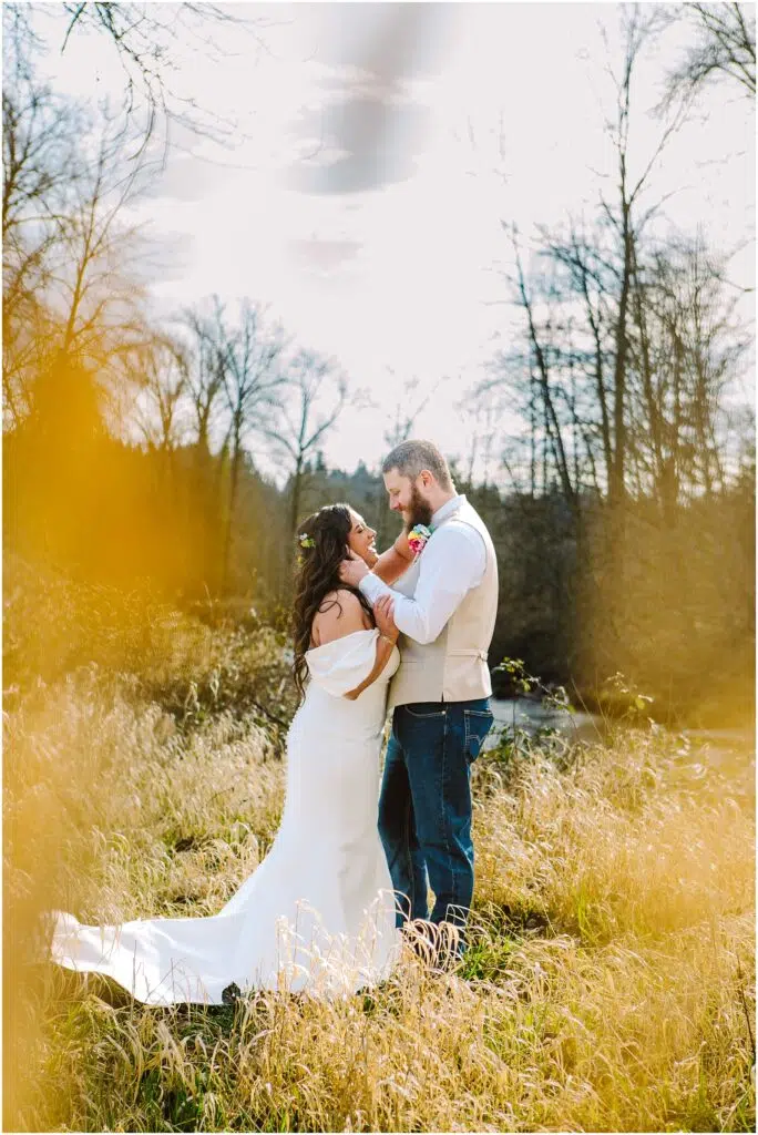 Snohomish Wedding Photographer GSquared Weddings A bride in a white dress and a groom in casual attire stand lovingly in a sunlit, grassy field near Machias. They are surrounded by tall trees and framed by soft, blurred foliage. The groom adjusts the brides hair, creating an intimate and tender moment. Serving Seattle, Snohomish and Washington State