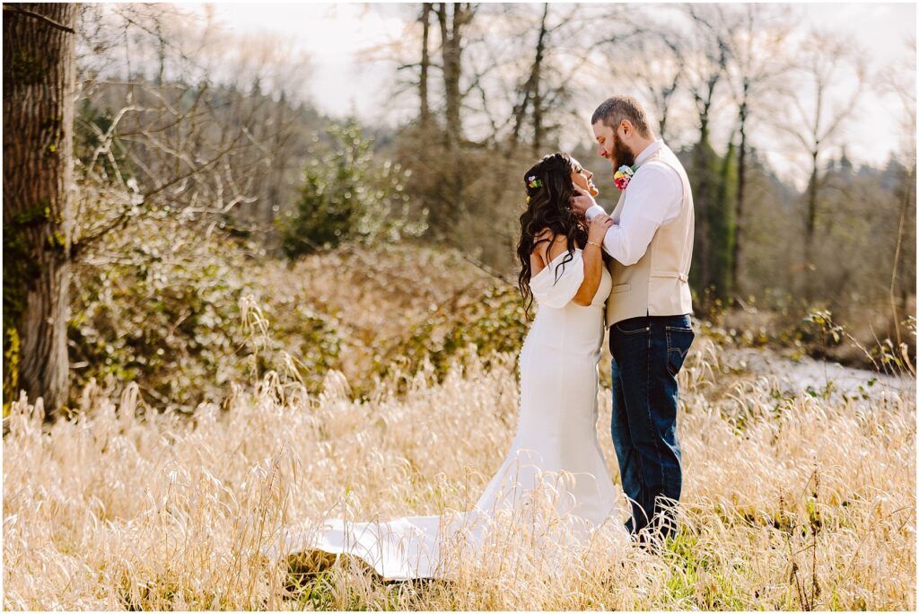 Snohomish Wedding Photographer GSquared Weddings In a field near Machias, the bride in a white off-the-shoulder dress and the groom in a vest and jeans stand amidst tall grasses. With trees as their backdrop, they face each other closely, creating an enchanting and romantic scene. Serving Seattle, Snohomish and Washington State