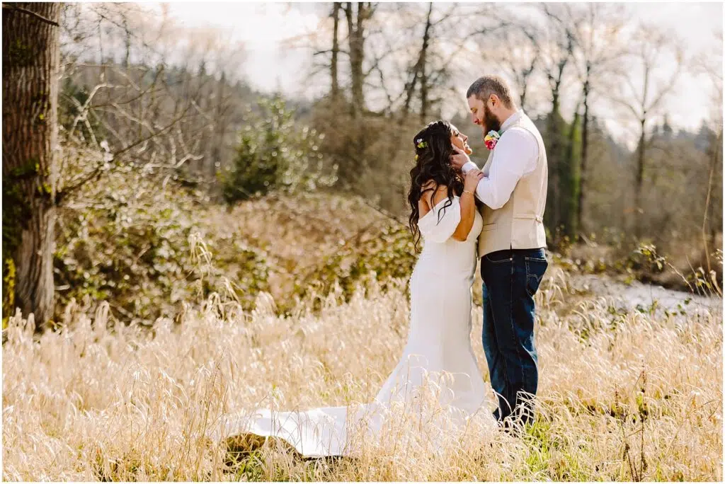 Snohomish Wedding Photographer GSquared Weddings In a field near Machias, the bride in a white off-the-shoulder dress and the groom in a vest and jeans stand amidst tall grasses. With trees as their backdrop, they face each other closely, creating an enchanting and romantic scene. Serving Seattle, Snohomish and Washington State