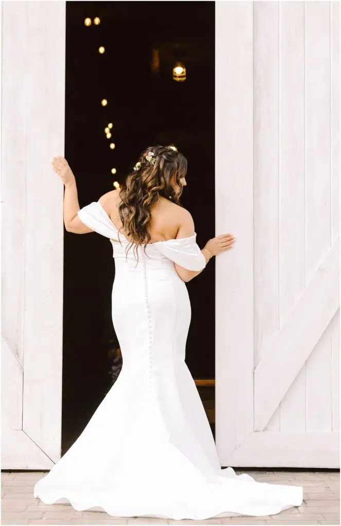 Snohomish Wedding Photographer GSquared Weddings A bride in an offshoulder white gown stands facing a large open barn door decorated with string lights Her long hair is styled with flowers and she looks back slightly in the serene Machias countryside highlighting the gowns button details and flowing train Serving Seattle Snohomish and Washington State Snohomish Wedding Photographer GSquared Weddings A bride in an off-shoulder white gown stands facing a large, open barn door decorated with string lights. Her long hair is styled with flowers, and she looks back slightly in the serene Machias countryside, highlighting the gowns button details and flowing train. Serving Seattle, Snohomish and Washington State