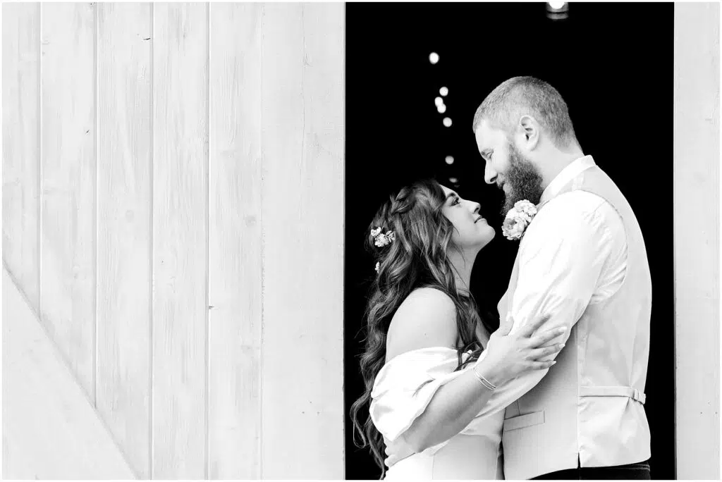 Snohomish Wedding Photographer GSquared Weddings A couple embraces lovingly in a Machias doorway. The woman looks up at the man, smiling. Both are dressed in wedding attire, with the man wearing a vest and the woman having flowers in her hair. The scene is beautifully captured in black and white. Serving Seattle, Snohomish and Washington State