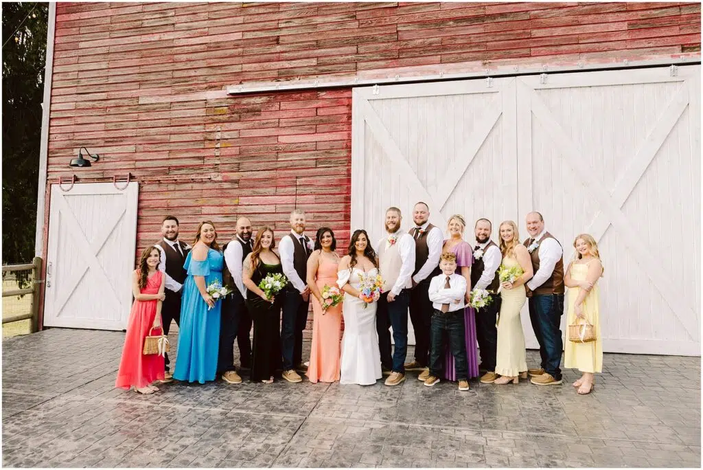 Snohomish Wedding Photographer GSquared Weddings A wedding party gathers joyfully in front of a charming red barn with large white doors, reminiscent of quaint Machias architecture. The group, dressed in formal attire, includes men in vests and women in colorful dresses. A young boy and girl are also part of the cheerful group, all smiling radiantly. Serving Seattle, Snohomish and Washington State