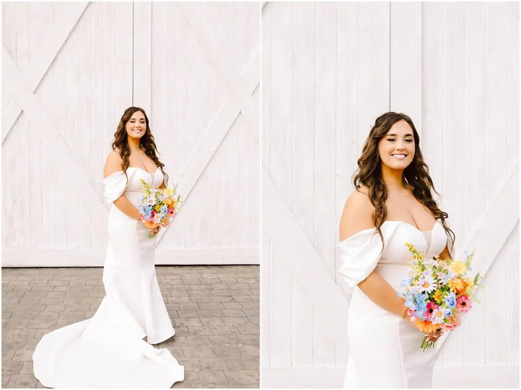 Snohomish Wedding Photographer GSquared Weddings Amidst a rustic white wooden backdrop in Machias, the bride dazzles in an elegant white gown. She clutches a vibrant bouquet of colorful flowers, her long, wavy hair cascading over her shoulders. The dress boasts off-the-shoulder sleeves and a sweeping train as she smiles serenely. Serving Seattle, Snohomish and Washington State