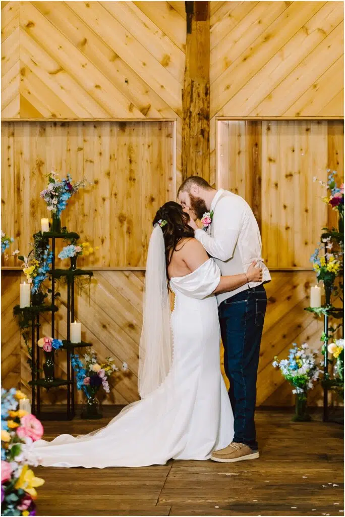 Snohomish Wedding Photographer GSquared Weddings In a rustic Machias setting, a couple shares a kiss among wooden walls and colorful floral arrangements. The bride wears a white gown and veil, while the groom sports a white shirt and jeans. Candles and vibrant flowers enhance the romantic atmosphere. Serving Seattle, Snohomish and Washington State
