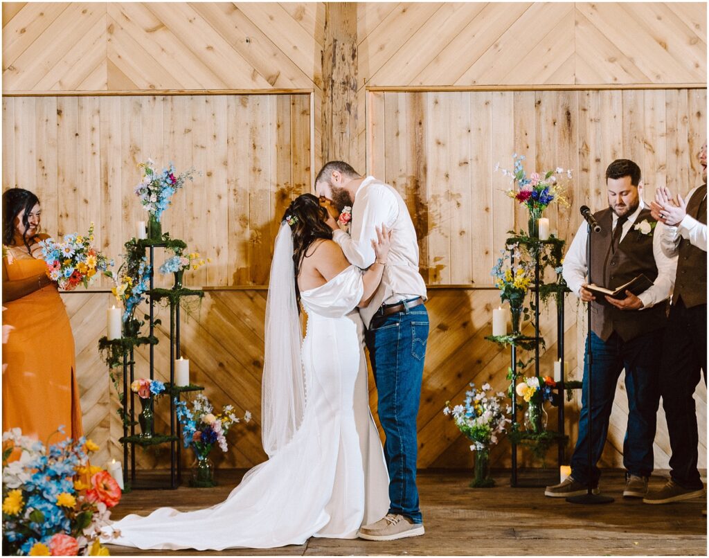 Snohomish Wedding Photographer GSquared Weddings A bride and groom share a kiss during their Machias wedding ceremony in front of a wooden backdrop. The groom is wearing jeans and a white shirt; the bride is in an off-shoulder white dress. They are surrounded by colorful flowers and guests clapping. Serving Seattle, Snohomish and Washington State
