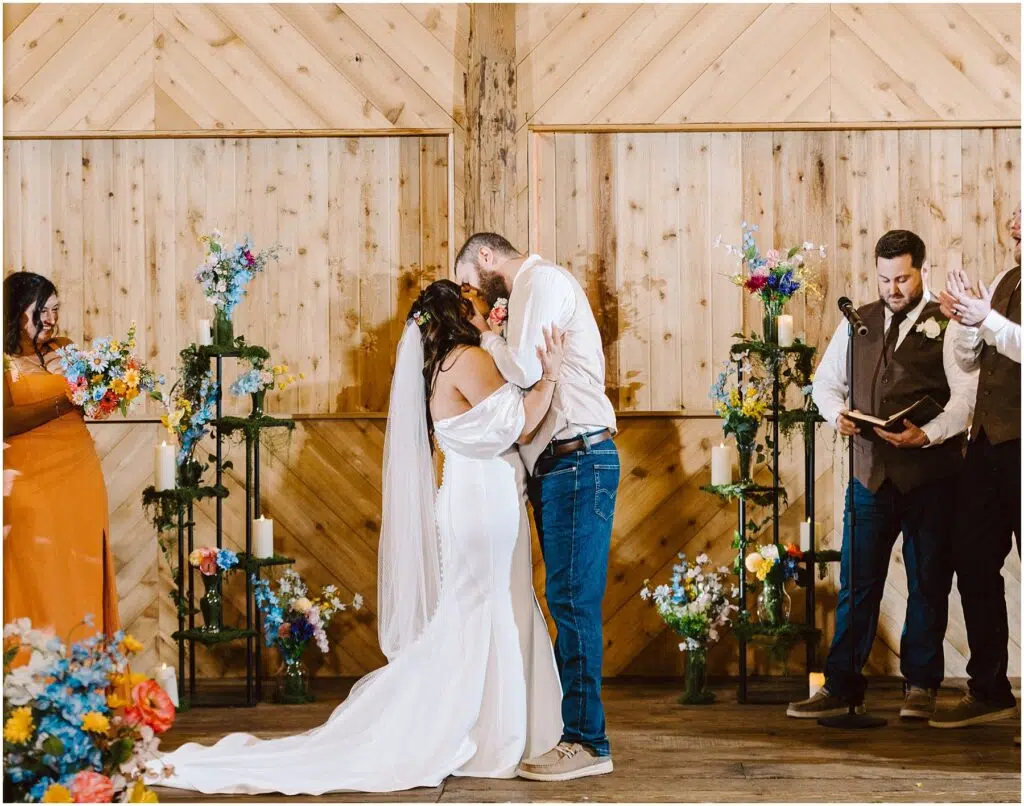 Snohomish Wedding Photographer GSquared Weddings A bride and groom share a kiss during their Machias wedding ceremony in front of a wooden backdrop. The groom is wearing jeans and a white shirt; the bride is in an off-shoulder white dress. They are surrounded by colorful flowers and guests clapping. Serving Seattle, Snohomish and Washington State