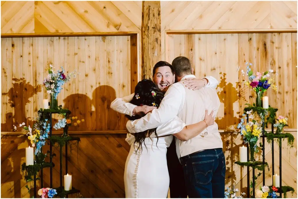 Snohomish Wedding Photographer GSquared Weddings Three people in a group hug at a wedding, standing in front of a wooden Machias wall. Flowers and candles decorate the surroundings, casting a warm glow. The person in the middle, wearing a vest, has a joyful expression. Serving Seattle, Snohomish and Washington State