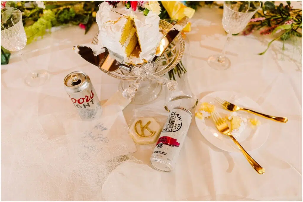 Snohomish Wedding Photographer GSquared Weddings A table in Machias showcases a sliced white frosted cake on a glass stand, flanked by a Coors Light can, a White Claw can, champagne flutes, a plate with cake, and gold utensils. Decorative flowers and greenery provide an elegant backdrop. Serving Seattle, Snohomish and Washington State