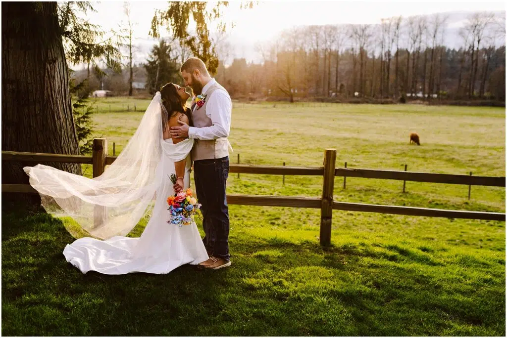 Snohomish Wedding Photographer GSquared Weddings A bride and groom embrace in a grassy field during a Machias sunset The brides veil flows in the breeze as she holds a colorful bouquet A wooden fence and trees frame the scene with a cow grazing peacefully in the distance Serving Seattle Snohomish and Washington State Snohomish Wedding Photographer GSquared Weddings A bride and groom embrace in a grassy field during a Machias sunset. The brides veil flows in the breeze as she holds a colorful bouquet. A wooden fence and trees frame the scene, with a cow grazing peacefully in the distance. Serving Seattle, Snohomish and Washington State