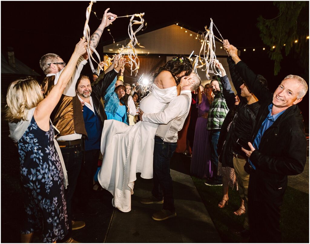 Snohomish Wedding Photographer GSquared Weddings Amid the joyful crowd in Machias, friends wave ribbon wands under the night sky. The man lifts the woman triumphantly as guests form a festive arch with ribbons, their warm laughter blending seamlessly with the celebration. Serving Seattle, Snohomish and Washington State