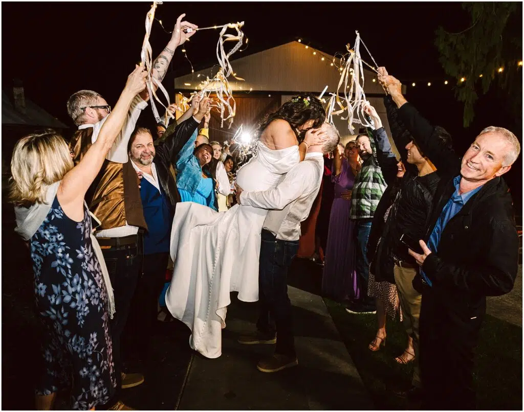 Snohomish Wedding Photographer GSquared Weddings Amid the joyful crowd in Machias, friends wave ribbon wands under the night sky. The man lifts the woman triumphantly as guests form a festive arch with ribbons, their warm laughter blending seamlessly with the celebration. Serving Seattle, Snohomish and Washington State