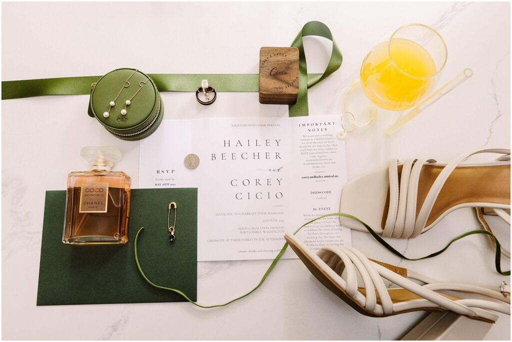 Snohomish Wedding Photographer GSquared Weddings Invitation set for Hailey Beecher and Corey Cicio features delicate perfume, green ribbon, pearl earrings, and a wooden item. An orange drink with a stirring stick complements this Port Gamble affair. Perfect touches for a Summer Wedding; RSVP and important notes included. Serving Seattle, Snohomish and Washington State