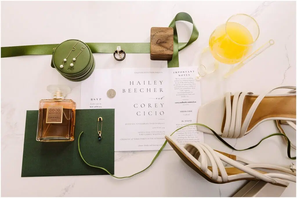 Snohomish Wedding Photographer GSquared Weddings Invitation set for Hailey Beecher and Corey Cicio features delicate perfume, green ribbon, pearl earrings, and a wooden item. An orange drink with a stirring stick complements this Port Gamble affair. Perfect touches for a Summer Wedding; RSVP and important notes included. Serving Seattle, Snohomish and Washington State