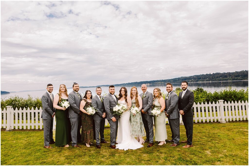 Snohomish wedding photographer gsquared weddings a wedding party poses in front of a white picket fence in port gamble with a scenic lake and forested hills as the backdrop Dressed in formal attire and holding flowers the group stands on the grassy area The sky is cloudy capturing the essence of a summer wedding Serving seattle snohomish and washington state Snohomish wedding photographer gsquared weddings a wedding party poses in front of a white picket fence in port gamble with a scenic lake and forested hills as the backdrop Dressed in formal attire and holding flowers the group stands on the grassy area The sky is cloudy capturing the essence of a summer wedding Serving seattle snohomish and washington state