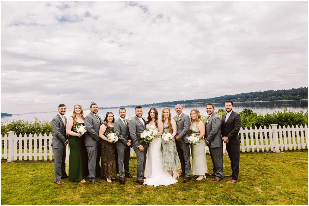 Snohomish Wedding Photographer GSquared Weddings A wedding party poses in front of a white picket fence in Port Gamble with a scenic lake and forested hills as the backdrop Dressed in formal attire and holding flowers the group stands on the grassy area The sky is cloudy capturing the essence of a summer wedding Serving Seattle Snohomish and Washington State Snohomish Wedding Photographer GSquared Weddings A wedding party poses in front of a white picket fence in Port Gamble, with a scenic lake and forested hills as the backdrop. Dressed in formal attire and holding flowers, the group stands on the grassy area. The sky is cloudy, capturing the essence of a summer wedding. Serving Seattle, Snohomish and Washington State