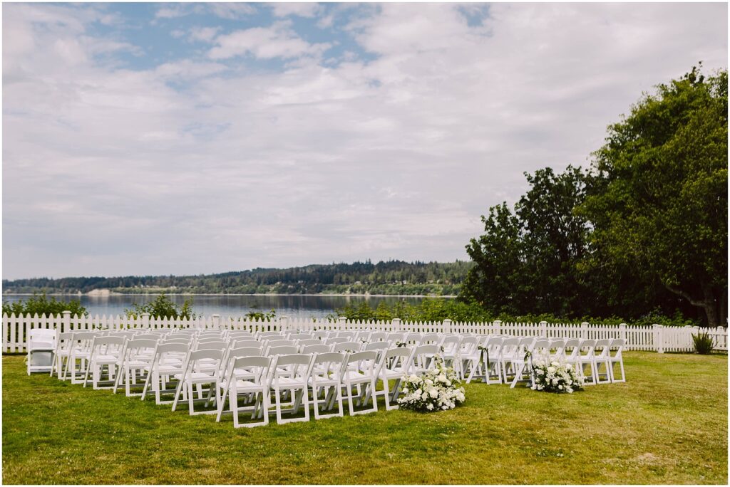 Snohomish wedding photographer gsquared weddings a serene summer wedding setup in port gamble features rows of white chairs arranged on a lush green lawn overlooking a calm lake White flower arrangements grace the aisles end while a white picket fence and trees stand proudly under the partly cloudy sky Serving seattle snohomish and washington state Snohomish wedding photographer gsquared weddings a serene summer wedding setup in port gamble features rows of white chairs arranged on a lush green lawn overlooking a calm lake White flower arrangements grace the aisles end while a white picket fence and trees stand proudly under the partly cloudy sky Serving seattle snohomish and washington state