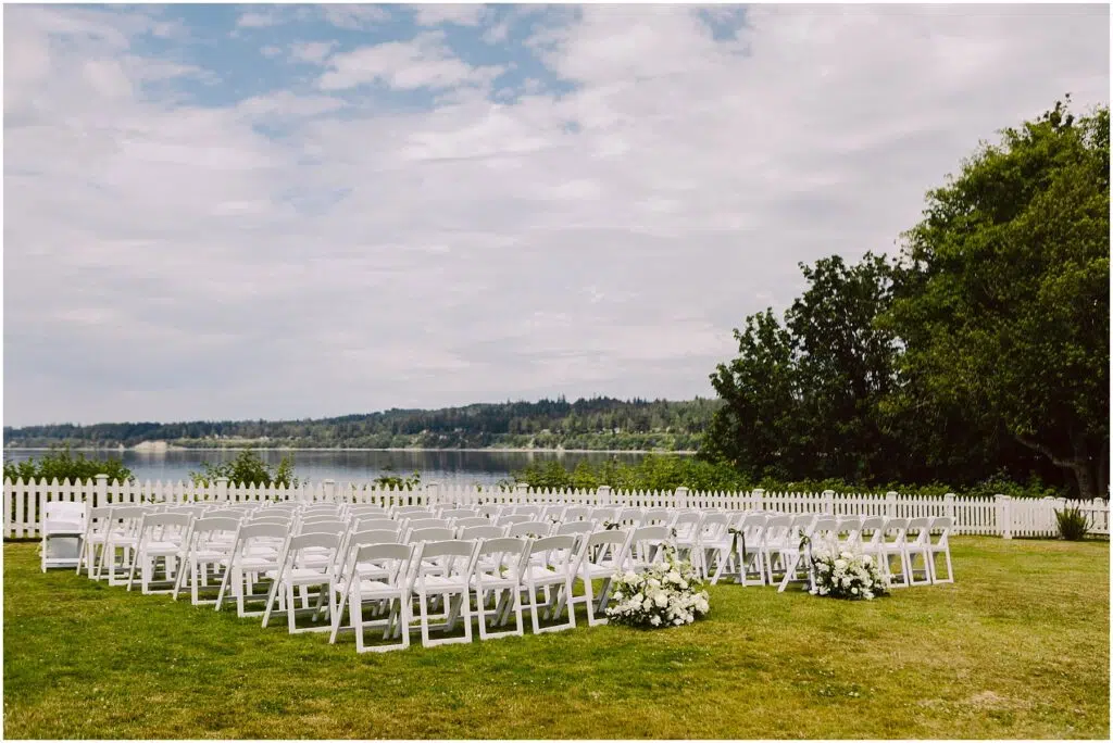 Snohomish Wedding Photographer GSquared Weddings A serene summer wedding setup in Port Gamble features rows of white chairs arranged on a lush green lawn overlooking a calm lake White flower arrangements grace the aisles end while a white picket fence and trees stand proudly under the partly cloudy sky Serving Seattle Snohomish and Washington State Snohomish Wedding Photographer GSquared Weddings A serene summer wedding setup in Port Gamble features rows of white chairs arranged on a lush green lawn, overlooking a calm lake. White flower arrangements grace the aisles end, while a white picket fence and trees stand proudly under the partly cloudy sky. Serving Seattle, Snohomish and Washington State
