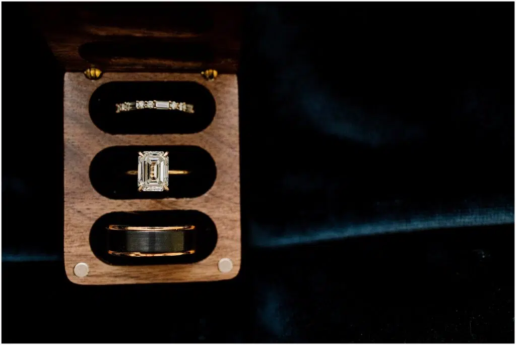 Snohomish Wedding Photographer GSquared Weddings A wooden box reveals three exquisite rings a thin diamond band on top a central emeraldcut diamond ring and a wider band below Perfect for a Port Gamble summer wedding the open box rests against a dark textured background that enhances their sparkle Serving Seattle Snohomish and Washington State Snohomish Wedding Photographer GSquared Weddings A wooden box reveals three exquisite rings: a thin diamond band on top, a central emerald-cut diamond ring, and a wider band below. Perfect for a Port Gamble summer wedding, the open box rests against a dark, textured background that enhances their sparkle. Serving Seattle, Snohomish and Washington State