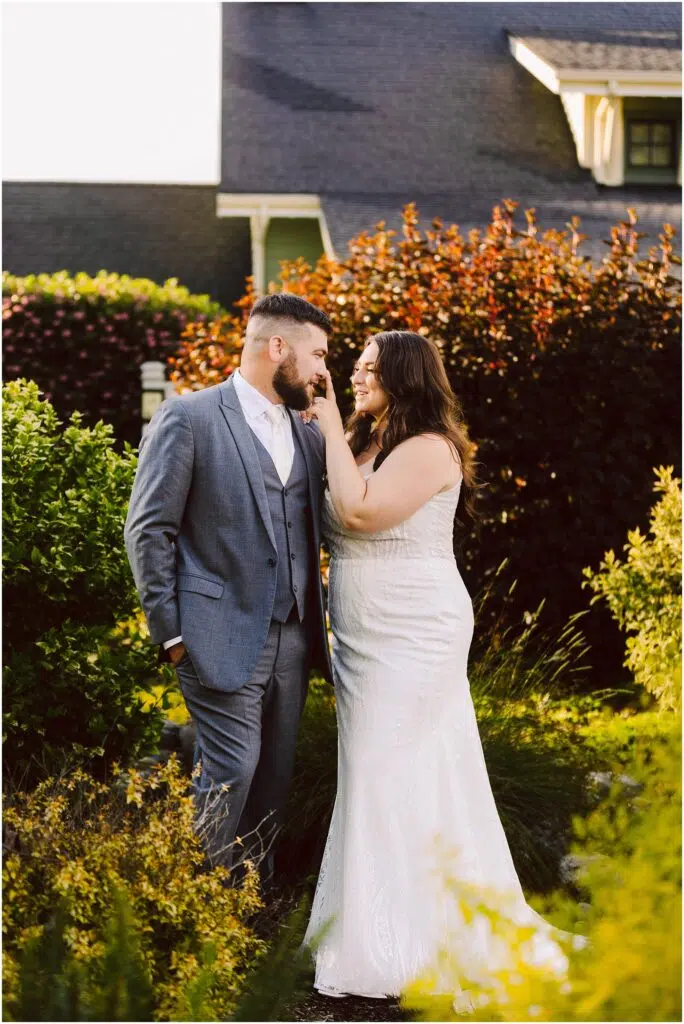 Snohomish Wedding Photographer GSquared Weddings A couple stands in a garden during their Port Gamble summer wedding the man in a grey suit and the woman in a white wedding dress She gently touches his beard as they smile at each other sunlight illuminating the greenery around them A charming house is visible in the background Serving Seattle Snohomish and Washington State Snohomish Wedding Photographer GSquared Weddings A couple stands in a garden during their Port Gamble summer wedding; the man in a grey suit and the woman in a white wedding dress. She gently touches his beard as they smile at each other, sunlight illuminating the greenery around them. A charming house is visible in the background. Serving Seattle, Snohomish and Washington State