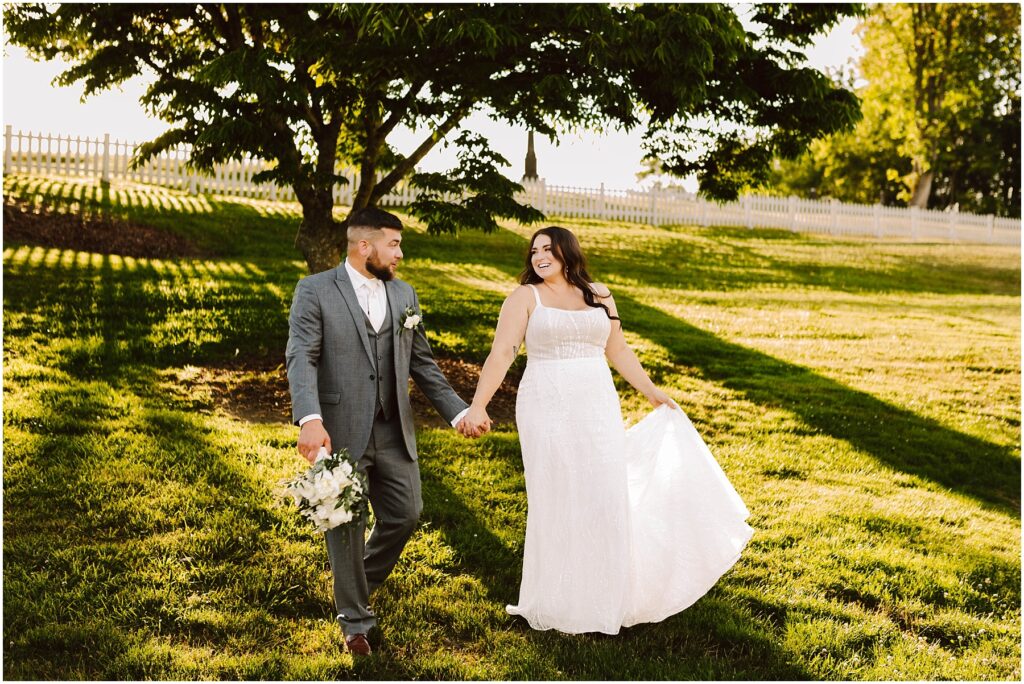 Snohomish wedding photographer gsquared weddings a smiling bride and groom hold hands strolling through a grassy field at their port gamble summer wedding The groom in a gray suit with a bouquet and the bride lifting her white dresss hem are framed by sunlit trees and a charming white fence in the background Serving seattle snohomish and washington state Snohomish wedding photographer gsquared weddings a smiling bride and groom hold hands strolling through a grassy field at their port gamble summer wedding The groom in a gray suit with a bouquet and the bride lifting her white dresss hem are framed by sunlit trees and a charming white fence in the background Serving seattle snohomish and washington state