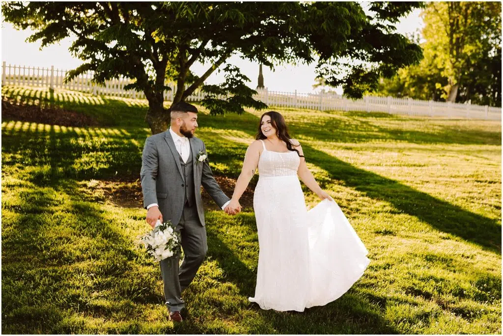 Snohomish Wedding Photographer GSquared Weddings A smiling bride and groom hold hands strolling through a grassy field at their Port Gamble summer wedding The groom in a gray suit with a bouquet and the bride lifting her white dresss hem are framed by sunlit trees and a charming white fence in the background Serving Seattle Snohomish and Washington State Snohomish Wedding Photographer GSquared Weddings A smiling bride and groom hold hands, strolling through a grassy field at their Port Gamble summer wedding. The groom, in a gray suit with a bouquet, and the bride, lifting her white dresss hem, are framed by sunlit trees and a charming white fence in the background. Serving Seattle, Snohomish and Washington State