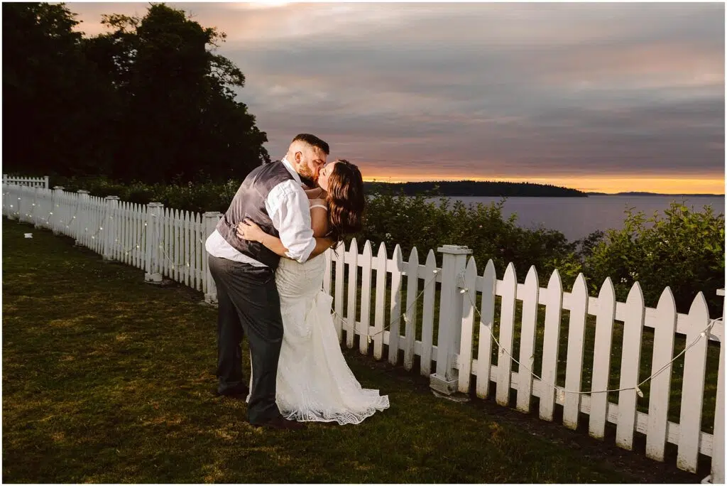 Snohomish Wedding Photographer GSquared Weddings A couple in wedding attire embraces and kisses beside a white picket fence at their summer wedding in Port Gamble with a picturesque lake and sunset in the background The evening sky displays shades of purple and orange Serving Seattle Snohomish and Washington State Snohomish Wedding Photographer GSquared Weddings A couple in wedding attire embraces and kisses beside a white picket fence at their summer wedding in Port Gamble, with a picturesque lake and sunset in the background. The evening sky displays shades of purple and orange. Serving Seattle, Snohomish and Washington State
