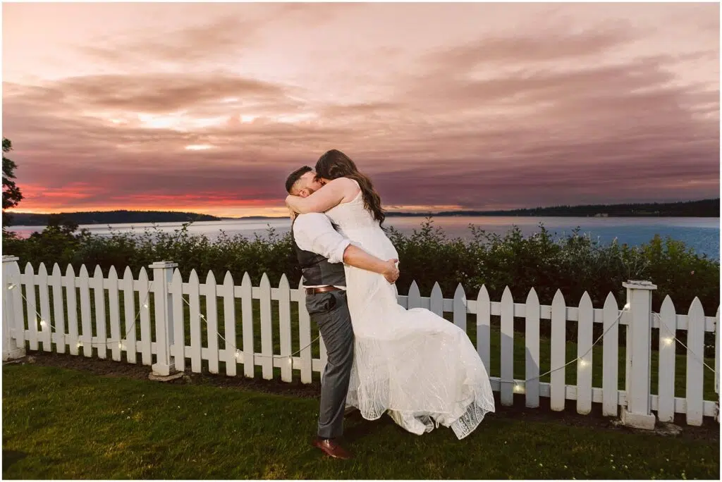 Snohomish Wedding Photographer GSquared Weddings A couple embraces joyfully in front of a white picket fence during sunset at their Summer Wedding The sky painted with hues of pink and orange over a calm lake in Port Gamble adds a romantic backdrop to their celebration The man lifts the woman who wears a white dress Serving Seattle Snohomish and Washington State Snohomish Wedding Photographer GSquared Weddings A couple embraces joyfully in front of a white picket fence during sunset at their Summer Wedding. The sky, painted with hues of pink and orange over a calm lake in Port Gamble, adds a romantic backdrop to their celebration. The man lifts the woman, who wears a white dress. Serving Seattle, Snohomish and Washington State