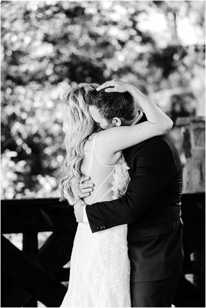 Snohomish Wedding Photographer GSquared Weddings A bride and groom embrace in a heartfelt hug amidst the scenic beauty of a winery. The bride, in an intricate lace gown with long hair cascading down her back, and the groom, in a dark suit, are framed by soft, blurred greenery. Black and white photo. Serving Seattle, Snohomish and Washington State