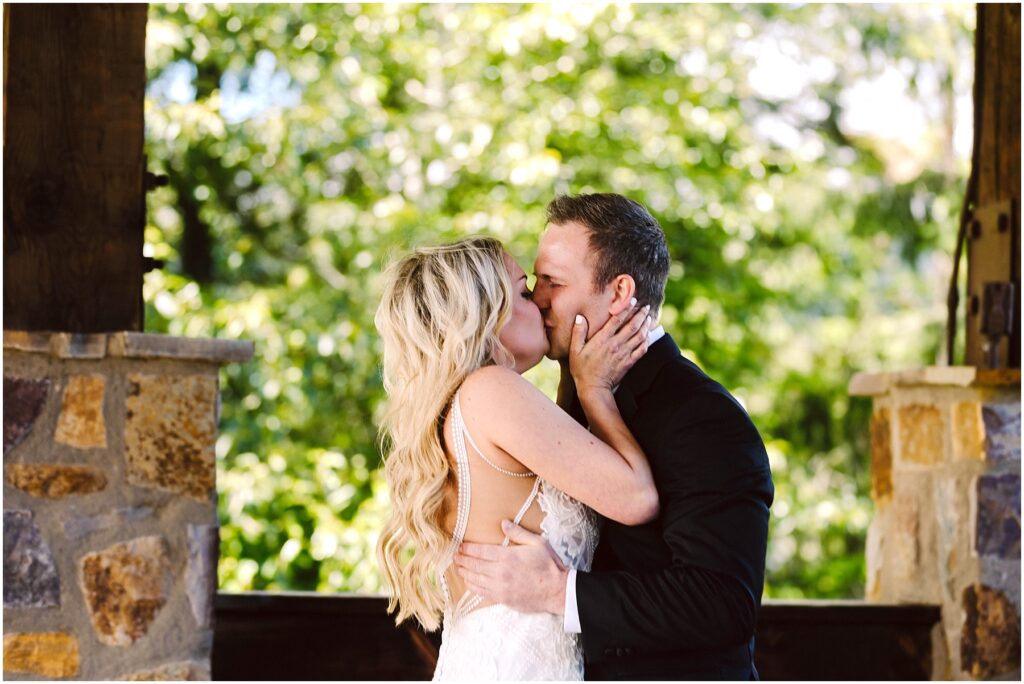 Snohomish Wedding Photographer GSquared Weddings A couple dressed in wedding attire shares a kiss under a stone and wood pavilion at a picturesque winery. The bride, with long blonde hair, and the groom in a black suit, are bathed in sunlight filtering through lush foliage, creating an enchanting and romantic setting. Serving Seattle, Snohomish and Washington State