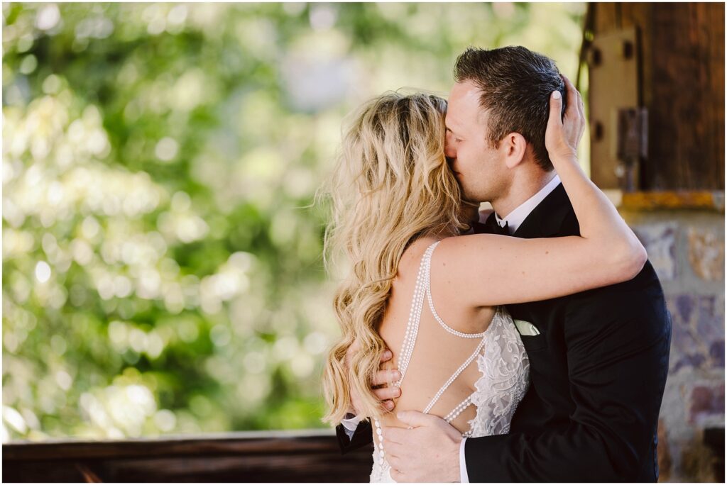Snohomish Wedding Photographer GSquared Weddings A couple in wedding attire shares a kiss outdoors at a charming winery. The groom, in a black suit, embraces the bride, who wears a white gown with lace details. Lush green foliage intermingles with the vineyard backdrop as the brides hand rests on the grooms shoulder. Serving Seattle, Snohomish and Washington State