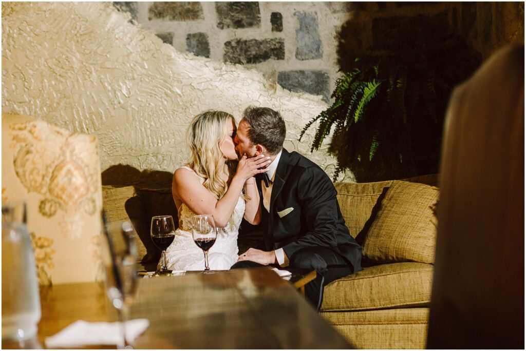 Snohomish Wedding Photographer GSquared Weddings A couple in formal attire kisses while seated on a couch, surrounded by the cozy ambiance of a rustic winery. A stone wall and lush plant provide the backdrop, as two glasses of red wine sit invitingly on the table before them, enhancing the intimate moment. Serving Seattle, Snohomish and Washington State