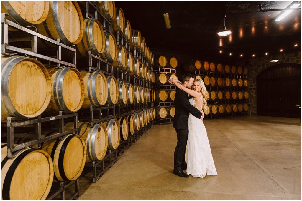 Snohomish Wedding Photographer GSquared Weddings A couple embraces while dancing in the winerys cellar, surrounded by stacks of wooden barrels. The woman is wearing a white dress, and the man is in a suit. Warm lighting creates a cozy atmosphere that perfectly complements the vineyards enchanting charm. Serving Seattle, Snohomish and Washington State
