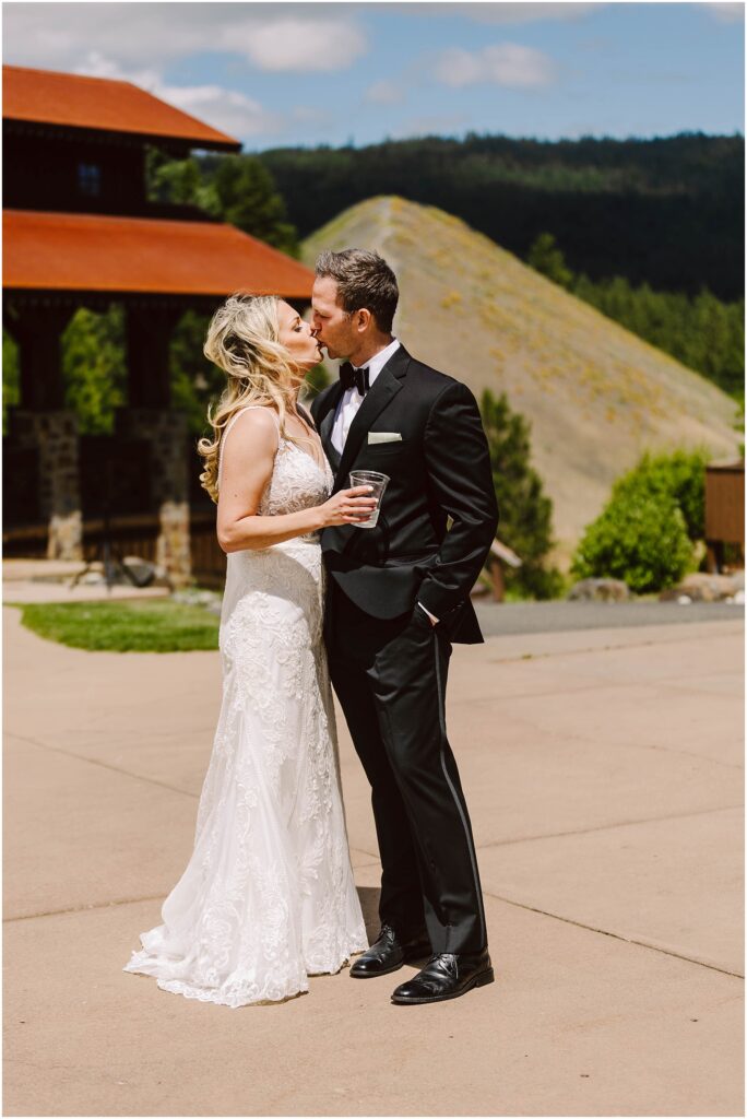 Snohomish Wedding Photographer GSquared Weddings A bride in a white lace dress and a groom in a black tuxedo kiss outdoors at a picturesque winery. The bride holds a drink as they stand on a paved area, with charming buildings and lush, hilly vineyard landscapes under the partly cloudy sky. Serving Seattle, Snohomish and Washington State