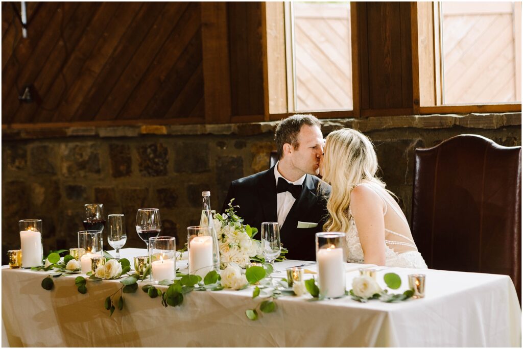 Snohomish Wedding Photographer GSquared Weddings A couple in formal attire kisses at a wedding reception table adorned with white flowers, candles, and greenery. They are seated in front of wooden walls and large windows of a charming winery, with wine glasses and a bottle gracing the table. Serving Seattle, Snohomish and Washington State