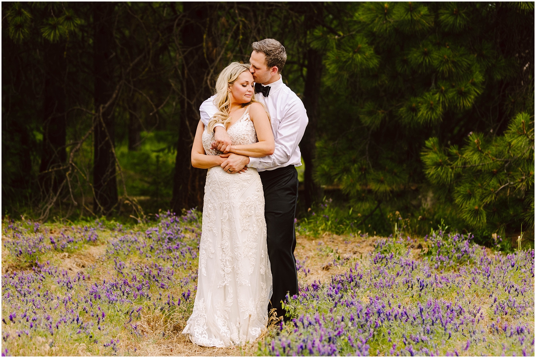 Snohomish Wedding Photographer GSquared Weddings A couple stands in a field of purple flowers near a quaint winery, surrounded by trees. The man, in a white shirt and black pants, embraces the woman, who wears a white lace dress. They both appear serene and content. Serving Seattle, Snohomish and Washington State