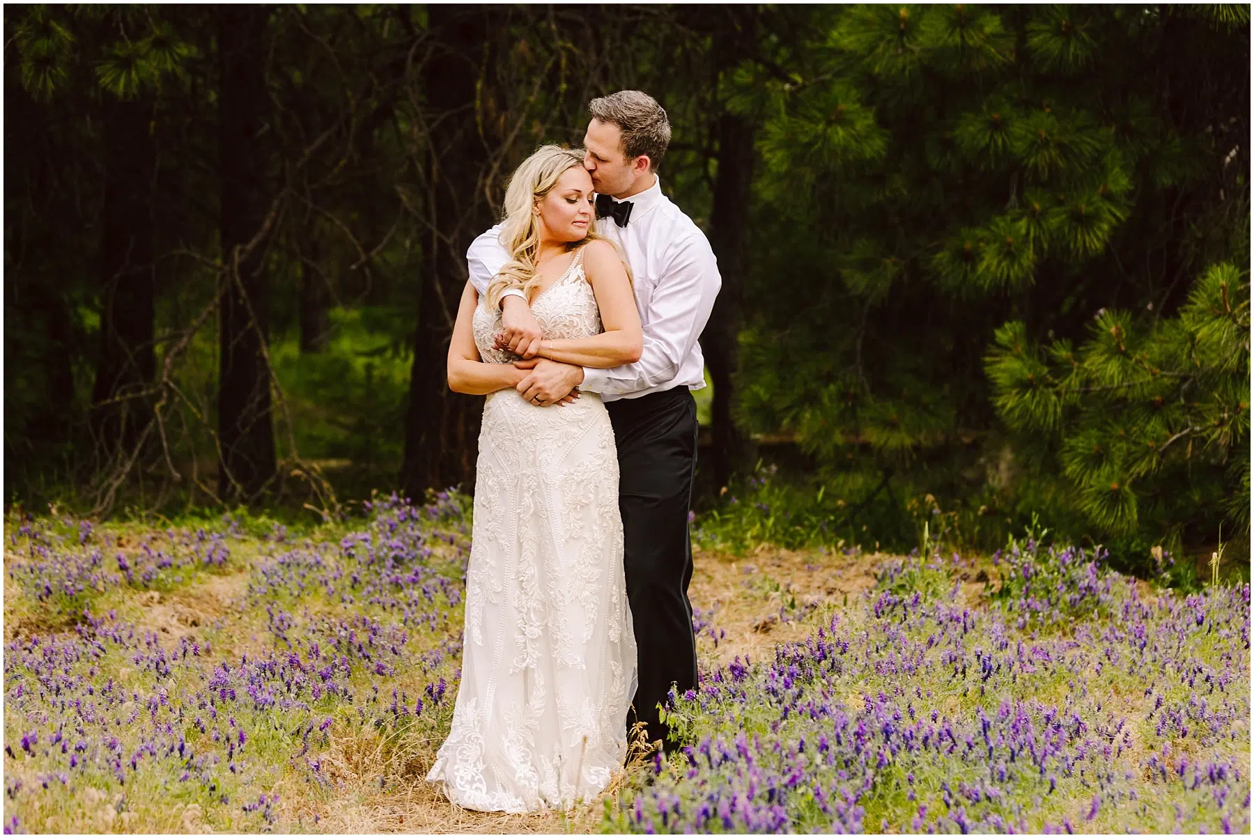 Snohomish Wedding Photographer GSquared Weddings A couple stands in a field of purple flowers near a quaint winery, surrounded by trees. The man, in a white shirt and black pants, embraces the woman, who wears a white lace dress. They both appear serene and content. Serving Seattle, Snohomish and Orlando