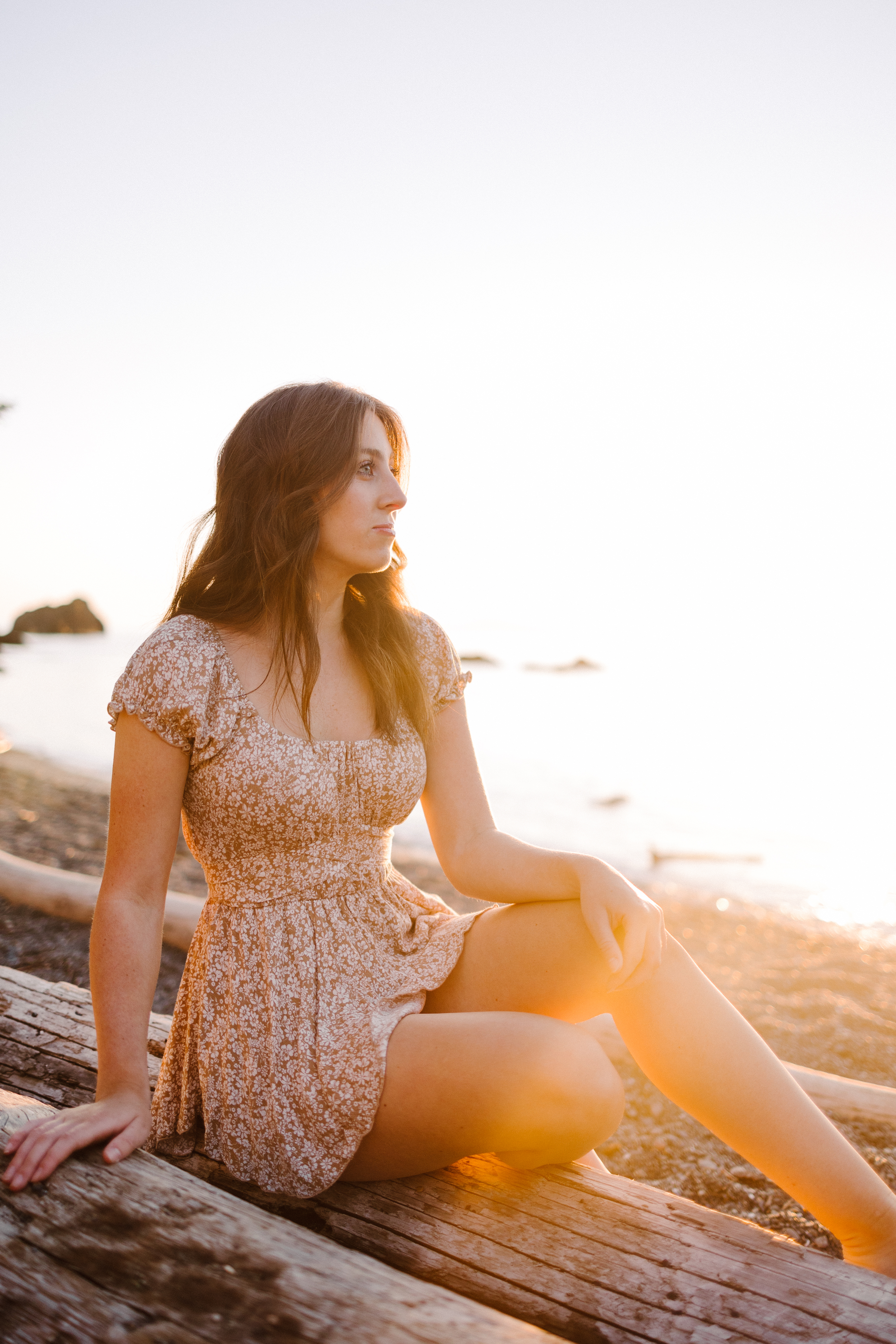 High school senior in a floral dress sitting on driftwood at the beach during golden hour High school senior in a floral dress sitting on driftwood at the beach during golden hour