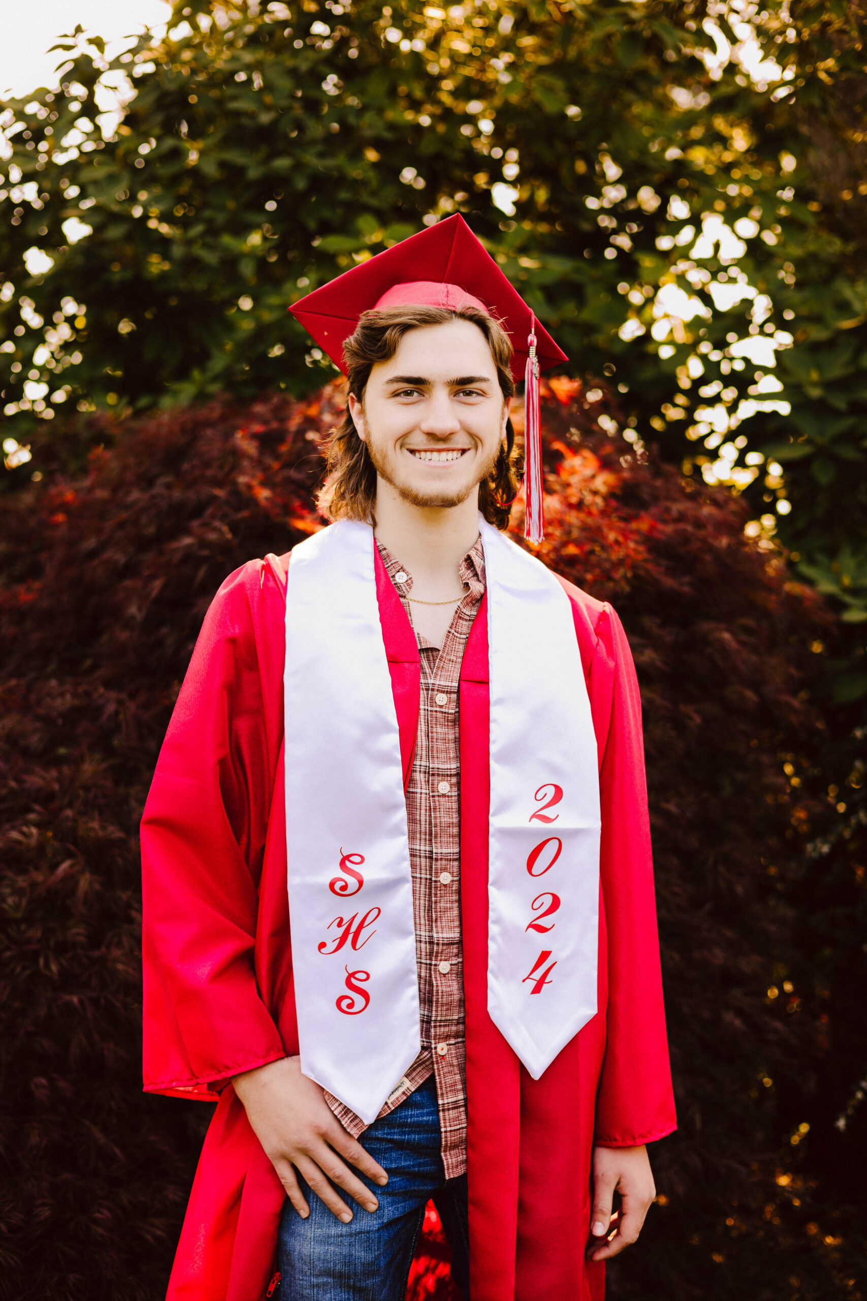 Snohomish wedding photographer gsquared weddings a gsquared senior stands outdoors radiating joy in a red graduation cap and gown They wear a white stole with red lettering shs 2024 over a plaid shirt Sunlight filters through the green foliage adding warmth to the celebration of their achievements Serving seattle snohomish and washington state