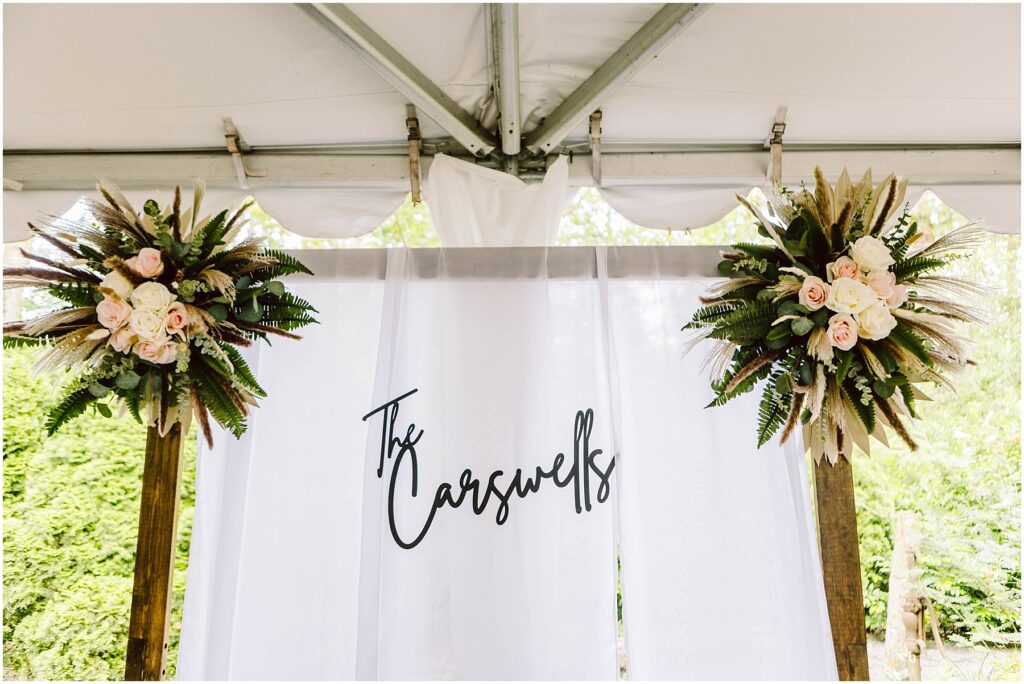 Snohomish Wedding Photographer GSquared Weddings A white wedding backdrop with the text The Carswells in elegant script stands gracefully in a Snohomish setting Flanking the backdrop are two floral arrangements featuring pink roses greenery and dried palm leaves all under a tent Serving Seattle Snohomish and Washington State Snohomish Wedding Photographer GSquared Weddings A white wedding backdrop with the text The Carswells in elegant script stands gracefully in a Snohomish setting. Flanking the backdrop are two floral arrangements featuring pink roses, greenery, and dried palm leaves, all under a tent. Serving Seattle, Snohomish and Washington State