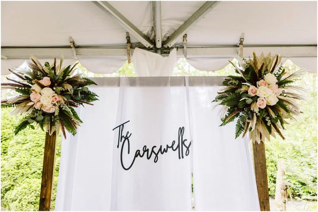 Snohomish Wedding Photographer GSquared Weddings A white wedding backdrop with the text The Carswells in elegant script stands gracefully in a Snohomish setting Flanking the backdrop are two floral arrangements featuring pink roses greenery and dried palm leaves all under a tent Serving Seattle Snohomish and Washington State Snohomish Wedding Photographer GSquared Weddings A white wedding backdrop with the text The Carswells in elegant script stands gracefully in a Snohomish setting. Flanking the backdrop are two floral arrangements featuring pink roses, greenery, and dried palm leaves, all under a tent. Serving Seattle, Snohomish and Washington State